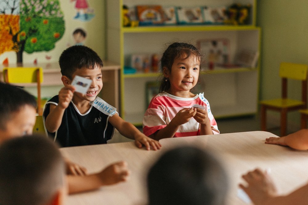 A boy and a girl at a classroom table, both smiling and holding up small picture cards during a game, other children seated around the table