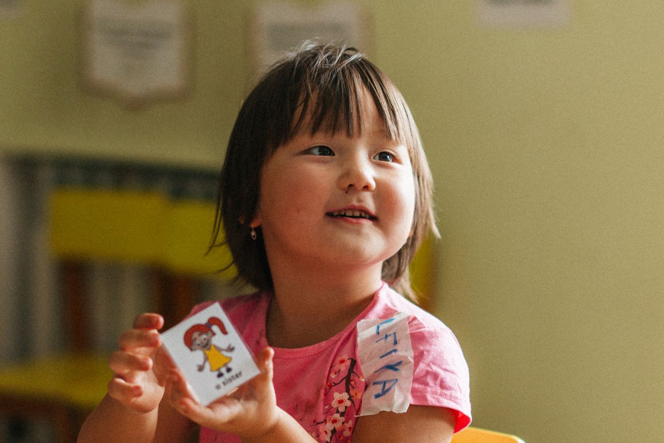 A girl holding up a small "a sister" flashcard with both hands, mouth open mid-word as she speaks